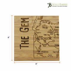 The 4-Piece Idaho Puzzle Coaster Set with Case by Totally Bamboo showcases a square bamboo coaster adorned with a laser-engraved map of Idaho, featuring the text "THE GEM" vertically. The distinctive design highlights cities such as Boise, Twin Falls, and Coeur d'Alene, along with notable landmarks and rivers. The "Totally Bamboo" logo is elegantly placed in the top right corner.