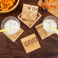 Two drinks with lemon slices on ice rest on a unique 4-Piece Idaho Puzzle Coaster Set, featuring laser-engraved maps and text like "The Gem State," from Totally Bamboo. A wooden tray, more of these intricately designed coasters, snacks, and a cheese platter adorn the dark wooden table.