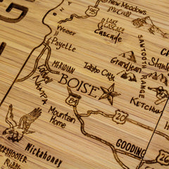 A close-up of the Totally Bamboo 4-Piece Idaho Puzzle Coaster Set with Case showcases a laser-engraved wooden map highlighting parts of Idaho, USA. It features cities such as Boise and Mountain Home, along with highways 84 and 20. The coaster set also includes illustrations of mountains and wildlife, like an eagle—perfect for a unique gift or home decor item.