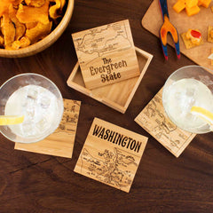 The 4-Piece Washington Puzzle Coaster Set with Case by Totally Bamboo, featuring intricately laser-engraved maps of Washington State and "The Evergreen State," is elegantly displayed on a table beneath two drinks garnished with lemon slices. Nearby, a bowl of chips and a tray with cheese and crackers complete this distinctive coaster setting.