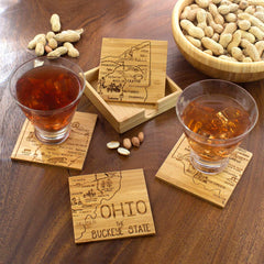 Two glasses filled with amber liquid sit on a 4-Piece Ohio Puzzle Coaster Set with Case by Totally Bamboo, featuring Ohio maps. They are surrounded by a wooden bowl of peanuts and more scattered peanuts, all placed on a rustic wooden surface.