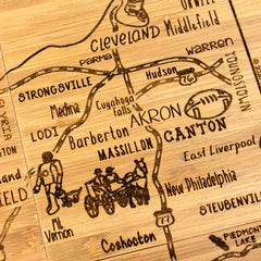 This laser-engraved artwork is a vintage-style map on wood depicting Ohio's cities, including Akron, Canton, and Cleveland. It showcases a football near Canton and cyclists near Barberton and Massillon. Crafted by Totally Bamboo, the 4-Piece Ohio Puzzle Coaster Set with Case is perfect for those in search of unique coasters.