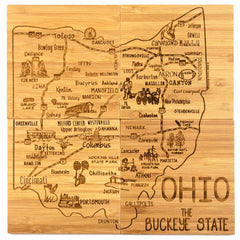 A wooden cutting board, laser-engraved with a map of Ohio highlighting cities such as Columbus, Cleveland, Cincinnati, and Toledo and embellished with local landmarks and symbols labeled "Ohio" and "The Buckeye State," mirrors the artistry in the 4-Piece Ohio Puzzle Coaster Set with Case from Totally Bamboo.