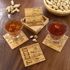 Two glasses of iced tea rest on laser-engraved wooden coasters from the Totally Bamboo 4-Piece Arizona Puzzle Coaster Set with Case, each displaying Arizona maps. A wooden box labeled "The Grand Canyon State" contains additional distinctive coasters. Nearby, a bowl of peanuts and scattered nuts lie on the wooden table, creating a cozy setting.