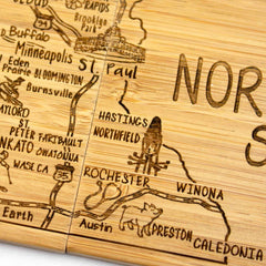 This 4-piece Minnesota Puzzle Coaster Set with Case from Totally Bamboo features laser-engraved artwork depicting a wooden map of southeastern Minnesota. It showcases cities such as Minneapolis, St. Paul, Rochester, Winona, and Austin and highlights local landmarks and wildlife with charming illustrations like a fish and a beaver—perfect for gifting on any occasion.