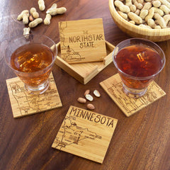 Two glasses with iced drinks sit atop laser-engraved wooden coasters displaying a map of Minnesota from the 4-Piece Minnesota Puzzle Coaster Set with Case by Totally Bamboo. A matching coaster stand labeled "The Northstar State" enhances the arrangement, making it ideal for gifting. A bowl of peanuts adds the finishing touch to the scene on the wooden table.