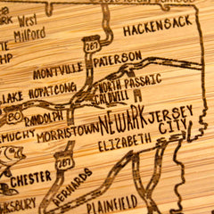 A 4-Piece New Jersey Puzzle Coaster Set with Case from Totally Bamboo features intricately carved wooden pieces, showcasing areas of New Jersey, including cities like Newark and Hackensack. The laser-engraved details highlight roads and smaller towns, making it a unique gift that offers a distinctive charm reminiscent of a carved wooden map.