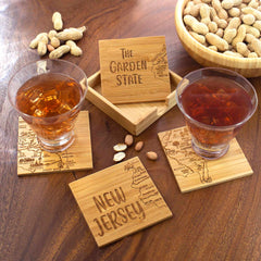 Two whiskey glasses are perched on the Totally Bamboo 4-Piece New Jersey Puzzle Coaster Set with Case, each coaster beautifully laser-engraved with New Jersey maps. One coaster reads "The Garden State" and another "New Jersey." A small bowl of peanuts and scattered peanuts add to the wooden table's charm, making these distinctive gifts an inviting touch to any gathering.