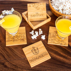 Two yellow drinks sit atop Totally Bamboo's 4-Piece Massachusetts Puzzle Coaster Set, each coaster uniquely laser-engraved with landmarks synonymous with the state. These coasters are placed on a wooden table next to a small case marked "The Bay State" and accompanied by a bowl of popcorn, creating a charming scene.