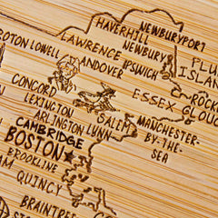 This intricately laser-engraved 4-Piece Massachusetts Puzzle Coaster Set with Case by Totally Bamboo features a beautifully detailed wooden map displaying the names of towns such as Boston, Cambridge, Lexington, and Salem. The design includes decorative illustrations that highlight places like Newburyport and Manchester-by-the-Sea, all elegantly carved into a light wood surface.