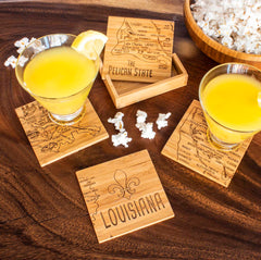 Two cocktails with lemon slices sit on distinctive coasters from the Totally Bamboo 4-Piece Louisiana Puzzle Coaster Set, each featuring detailed laser-engraved artwork of Louisiana maps. Close by, a wooden bowl of popcorn sits alongside an extra coaster from the same set, decorated with "Louisiana" and a fleur-de-lis motif.