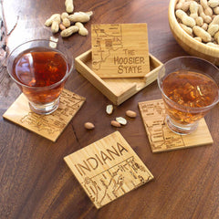 Two glasses of amber liquid sit on intricately laser-engraved coasters crafted by Totally Bamboo, each featuring a beautifully detailed map of Indiana and the phrase "The Hoosier State." This delightful 4-Piece Indiana Puzzle Coaster Set with Case is an ideal gift choice, presented alongside a wooden box and a bowl of peanuts on the brown wooden table.