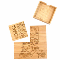 Totally Bamboo's 4-Piece Indiana Puzzle Coaster Set is spread out, showcasing individual wooden coasters that feature a laser-engraved map of Indiana with city names. One coaster stands upright, highlighting the state's outline with "Indiana" engraved. The set includes a wooden holder to complement this distinctive collection. A larger piece is etched with "The Hoosier State," making it an ideal gift choice.