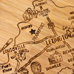 A close-up of the "4-Piece Kentucky Puzzle Coaster Set with Case" by Totally Bamboo showcases laser-engraved wooden details, highlighting cities such as Louisville, Lexington, and Frankfort. Near "Big Bone Lick State Park," an illustration of a mammoth is featured. Frankfort's location is marked with a star, adding character to this unique coaster set.