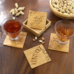 Two glasses with amber-colored drinks sit on the unique, laser-engraved 4-Piece Kentucky Puzzle Coaster Set by Totally Bamboo, featuring a map design and the text "KENTUCKY." Nearby, a wooden case labeled "THE BLUEGRASS STATE" rests on a table beside a bowl of peanuts.