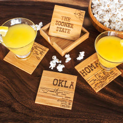 The 4-Piece Oklahoma Puzzle Coaster Set with Case by Totally Bamboo is showcased on a wooden table, supporting two yellow drinks with lemon wedges. To complement the scene, there's also a bowl of popcorn and scattered popcorn kernels.