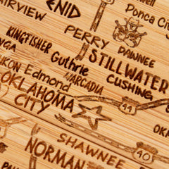 The Totally Bamboo 4-Piece Oklahoma Puzzle Coaster Set with Case showcases a close-up of a wooden map featuring carved names and locations in Oklahoma, including Perry, Guthrie, Stillwater, Cushing, Edmond, Norman, and Oklahoma City. This elegant laser-engraved set includes a star marking Oklahoma City for a unique touch.