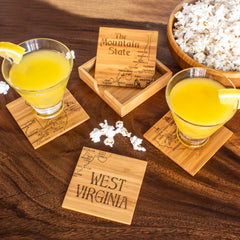 Two cocktails with lemon wedges rest on laser-engraved coasters, part of the 4-Piece West Virginia Puzzle Coaster Set with Case by Totally Bamboo. These coasters feature a map of West Virginia, labeled "The Mountain State." A matching case displaying the state puzzle coaster set and a bowl of popcorn complete the scene on a wooden table—ideal for unique coaster gifts.