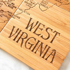 A laser-engraved wooden board from Totally Bamboo, resembling the 4-Piece West Virginia Puzzle Coaster Set with Case, displays a carved map outline with place names of "West Virginia." This unique coaster gift sits elegantly on a light surface.