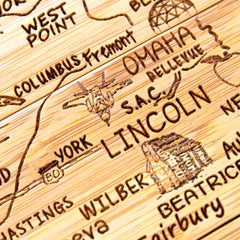 A close-up of a wooden map from the 4-Piece Nebraska Puzzle Coaster Set with Case by Totally Bamboo showcases Nebraska with engraved landmarks like Lincoln and Omaha. The laser-engraved details provide a textured appearance, beautifully highlighting towns such as York and Wilber.