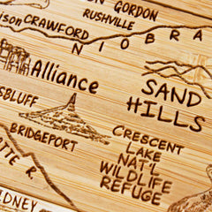 A detailed wooden carving from the Totally Bamboo 4-Piece Nebraska Puzzle Coaster Set with Case features a map highlighting locations such as Alliance, Sand Hills, Crescent Lake Nat'l Wildlife Refuge, and Bridgeport. The topographic bluffs are artistically laser-engraved into the wood, creating an exquisite gift set.