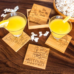 Two cocktails with lemon slices sit on wooden coasters from the 4-Piece Missouri Puzzle Coaster Set by Totally Bamboo, featuring a laser-engraved map and "The Show-Me State." Popcorn is scattered around and fills a bowl nearby, enhancing the cozy atmosphere.