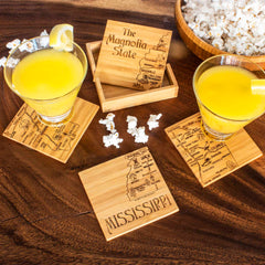 Two yellow drinks with lemon slices sit on distinctive State Puzzle Coasters by Totally Bamboo, featuring Mississippi maps and text. An open wooden box labeled "The Magnolia State" is nearby, accompanied by a wooden bowl of popcorn, all displayed on a wooden table adorned with laser-engraved artwork.