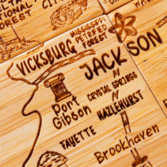 A decorative wooden map showcases laser-engraved artwork of locations like Vicksburg, Jackson, and the Mississippi Petrified Forest. This unique piece complements the precision of our 4-Piece Mississippi Puzzle Coaster Set with Case by Totally Bamboo, featuring landmarks such as Port Gibson, Crystal Springs, and Hazlehurst.