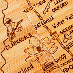 A detailed engraving on the artwork showcases a map featuring Clarksdale, Batesville, and Greenwood. Near Clarksdale, a musician with a guitar is skillfully etched, and below it lies an intricately carved magnolia flower. This design evokes the 4-Piece Mississippi Puzzle Coaster Set with Case by Totally Bamboo.