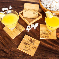 On a wooden table, two glasses of yellow drink garnished with lemon slices rest on exquisite coasters from the 4-Piece Maryland Puzzle Coaster Set with Case by Totally Bamboo. Nearby, a wooden dish filled with popcorn sits adjacent to another coaster highlighting the engraved text "MARYLAND Old Line State," emphasizing the distinctive artistry of this state-themed coaster set.