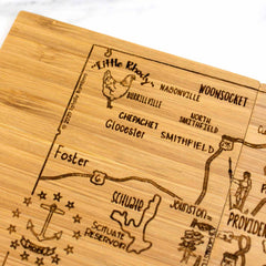 A wooden cutting board adorned with a map of various Rhode Island areas, such as Woonsocket, Burrillville, and North Smithfield, labeled "Little Rhody." The map includes illustrations of a rooster, an anchor, and a fish. It's an ideal addition to any kitchen when paired with the 4-Piece Rhode Island Puzzle Coaster Set with Case by Totally Bamboo.