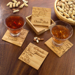 Two drinks rest on a distinctive 4-piece puzzle coaster set by Totally Bamboo, featuring laser-engraved maps of Rhode Island. A wooden case labeled "The Ocean State" sits open nearby, while a bowl of peanuts complements the scene on a rustic table.