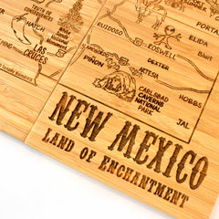 The 4-Piece New Mexico Puzzle Coaster Set with Case by Totally Bamboo offers a distinctive wooden map that functions as a state puzzle coaster. It highlights New Mexico with laser-engraved labels of cities and landmarks such as Roswell, Carlsbad Caverns National Park, and Las Cruces. The inscription at the bottom reads "New Mexico: Land of Enchantment.