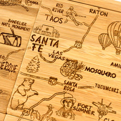 A close-up of the Totally Bamboo 4-Piece New Mexico Puzzle Coaster Set with Case displays intricately carved areas of New Mexico, featuring landmarks, roads, a skier, a bear, a hot air balloon, pine trees, and symbols for towns like Taos and Santa Fe.