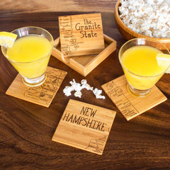 Two glasses of orange juice with lemon wedges rest on a Totally Bamboo 4-Piece New Hampshire Puzzle Coaster Set, which showcases laser-engraved maps and text about "The Granite State." Nearby on the wooden table is a bowl of popcorn, presenting a distinctive gift idea. The included coaster holder sits in the center.