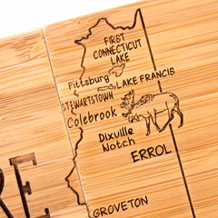 A wooden carving showcasing a map of northern New Hampshire with labeled towns, such as Pittsburg, Stewartstown, and Colebrook. It includes an illustration of a moose near Lake Francis and is part of the 4-Piece New Hampshire Puzzle Coaster Set with Case by Totally Bamboo—an ideal gift for nature enthusiasts.