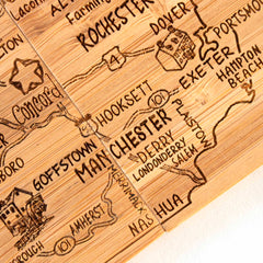 The 4-Piece New Hampshire Puzzle Coaster Set with Case by Totally Bamboo is a meticulously crafted wooden map that showcases towns like Manchester, Nashua, Concord, and Dover. This unique gift set features laser-engraved coasters as decorative elements, incorporating designs such as a compass rose and small etched drawings on the surface to highlight landmarks.