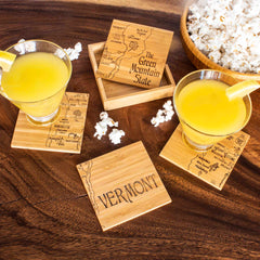 Two yellow cocktails garnished with lemon slices rest on the 4-Piece Vermont Puzzle Coaster Set by Totally Bamboo, each coaster adorned with maps of Vermont and "The Green Mountain State." A small wooden case and scattered popcorn are nearby on the wooden table, making perfect companions to this distinctive coaster set.