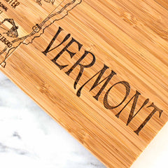 A close-up of a wooden board from the 4-Piece Vermont Puzzle Coaster Set with Case by Totally Bamboo showcases the word "Vermont" along with intricate illustrations and place names visible on its side. The board is elegantly displayed on a white marble surface.