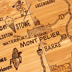 An intricate close-up showcases the 4-Piece Vermont Puzzle Coaster Set with Case by Totally Bamboo, featuring a wooden, laser-engraved map of Vermont. It highlights notable locations such as Montpelier, Barre, Stowe, and Greensboro accompanied by delightful illustrations of a skier, an ice cream cone, and corn. Highways 89 and 2 elegantly weave through this distinctive coaster collection.