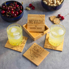 Two glasses with ice rest on distinctive coasters featuring maps of Nevada. At the center is a wooden box inscribed with "Nevada The Silver State." Nearby, bowls filled with cherries, peanuts, and crackers sit on a gray surface, perfectly complementing this charming 4-Piece Nevada Puzzle Coaster Set with Case from Totally Bamboo.