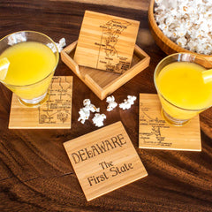 The table is elegantly adorned with the 4-Piece Delaware Puzzle Coaster Set with Case by Totally Bamboo, each coaster laser-engraved to highlight Delaware as "The First State." Two glasses of orange juice topped with lemon wedges rest on these distinctive coasters, while a tempting bowl of popcorn sits nearby.