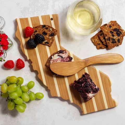 The California Serving Board with Spreader Knife Gift Set by Totally Bamboo features sliced bread, cheese, blackberries, and raspberries beautifully arranged alongside a glass of white wine. A bamboo spreader knife is elegantly placed amidst green grapes and vibrant red flowers, all set against a pristine white background.