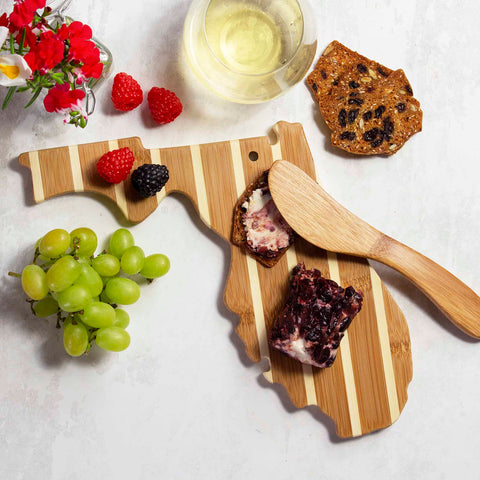 The Florida Serving Board with Spreader Knife Gift Set by Totally Bamboo elegantly displays crackers adorned with jam, accompanied by its bamboo spreader knife. Beside it, a glass of white wine, fresh green grapes, raspberries, blackberries, and a vase of flowers complement the perfect charcuterie setup on a pristine white surface.