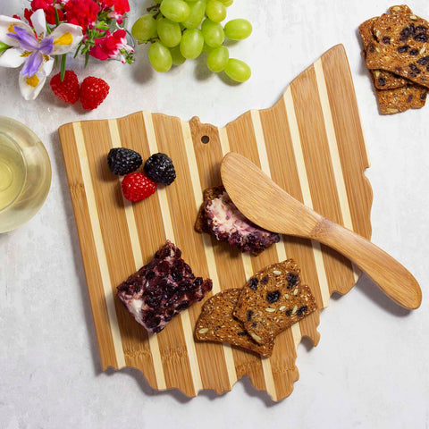 The Ohio Serving Board with Spreader Knife Gift Set by Totally Bamboo beautifully presents berry-topped cheese and crackers. Nearby, grapes and a glass of white wine enhance the setting, accented by fresh flowers and berries.