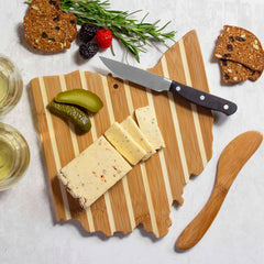 Cheese slices are elegantly arranged on the Ohio Serving Board with Spreader Knife Gift Set from Totally Bamboo, accompanied by pickles, berries, and crispbread. Two glasses of white wine enhance the scene, while a sprig of rosemary adds a charming touch.