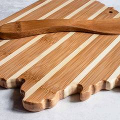 The Ohio Serving Board with Spreader Knife Gift Set by Totally Bamboo features a wooden charcuterie board designed with alternating light and dark stripes in the shape of the United States. This stylish set includes a matching spreader knife that complements the elegant display on a white marbled countertop.