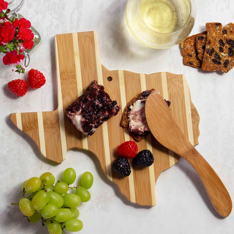 The Texas Serving Board with Spreader Knife Gift Set by Totally Bamboo showcases an artful arrangement of crackers, cheese, and berries, accompanied by a bamboo spreading knife. A glass of white wine perfectly complements the green grapes and vibrant red flowers beautifully displayed on a crisp white surface.