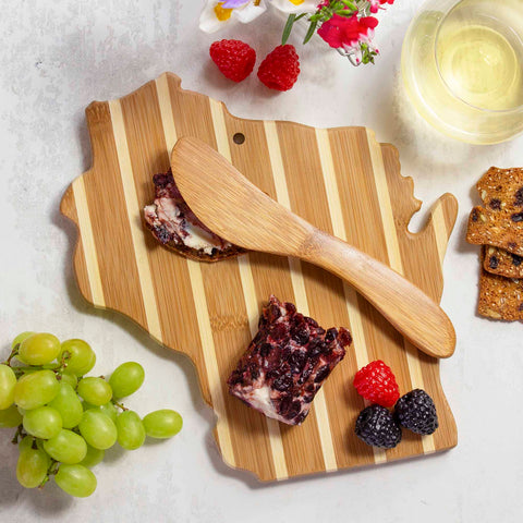 A Totally Bamboo Wisconsin Serving Board with Spreader Knife Gift Set showcases a piece of cheese topped with berries and accompanied by a bamboo spreading knife. Grapes, crackers, raspberries, blackberries, a glass of white wine, and flowers are elegantly arranged around the charcuterie board.