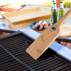 A Totally Bamboo BBQ Grill Scraper with Bottle Opener is used to clean a barbecue grill, as a wooden tray sits nearby. In the outdoor patio setting, there is also a plate with sausages and three open bottles of beer.